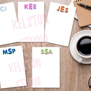 May include: A collection of personalized stationery pads with colorful initials. The pads are white with initials in blue, pink, orange, and green. A cup of coffee, glasses, a pen, and a notebook are on a wooden surface.