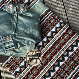 May include: A sage green shirt with decorative collar and cuffs, alongside a patterned blanket with red, white, and blue geometric designs. A circular emblem with a horse logo is also visible.