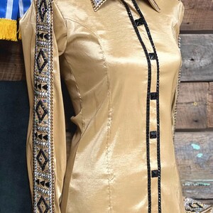 May include: A gold satin long-sleeve shirt with black trim and rhinestones. The shirt has a western-style collar and button closure.