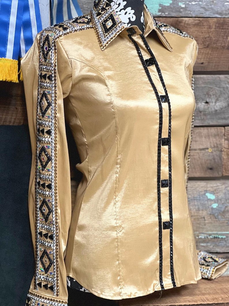 May include: A gold satin long-sleeve shirt with black trim and rhinestones. The shirt has a western-style collar and button closure.