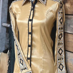 May include: A gold satin long-sleeve shirt with black trim and rhinestones. The shirt has a geometric pattern on the sleeves and collar.