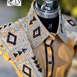 May include: A gold show shirt with black and silver embellishments. The shirt has a geometric pattern with rhinestones and black accents. The shirt is on a mannequin.