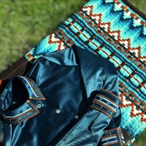 May include: Teal riding shirt with an embellished collar and cuffs, placed on a patterned saddle pad. The shirt has a satin finish and decorative buttons. The saddle pad features a geometric design in shades of blue, brown, and white.