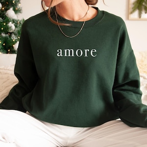 May include: A dark green sweatshirt with the word "amore" in white, centered on the chest. The long-sleeved top is made of a soft material. The sweatshirt is a comfortable and stylish piece of apparel.