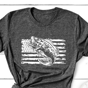 May include: Dark gray t-shirt featuring a white graphic of a fish leaping out of the water, set against a distressed American flag design. The shirt is styled with rolled sleeves and a knot at the side, with white sneakers and small potted plants nearby.