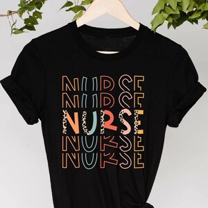 May include: A black t-shirt with the word 'NURSE' printed on it in a rainbow of colors. The word is repeated five times, with each repetition slightly offset from the one above it. The word 'NURSE' is also printed in a leopard print pattern in the center of the design.