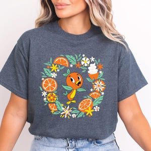 Puede incluir: Camiseta gris oscuro con un dise&ntilde;o de p&aacute;jaro naranja de dibujos animados, rodeado de rodajas de naranja, flores y un peque&ntilde;o postre. El gr&aacute;fico est&aacute; centrado, creando un dise&ntilde;o gr&aacute;fico colorido.