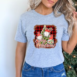 May include: A gray t-shirt with a Christmas-themed graphic featuring three gnomes standing around a Christmas tree. The graphic has a red and black buffalo plaid background and the text "Merry Christmas!"