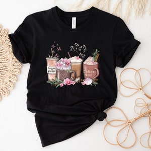 May include: Black t-shirt featuring a graphic of three coffee cups with whipped cream and flowers. The text on the cups reads "Coffee please", "Coffee first!" and a heart.