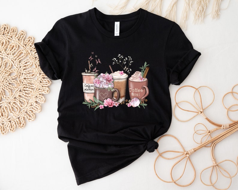 May include: Black t-shirt featuring a graphic of three coffee cups with whipped cream and flowers. The text on the cups reads "Coffee please", "Coffee first!" and a heart.