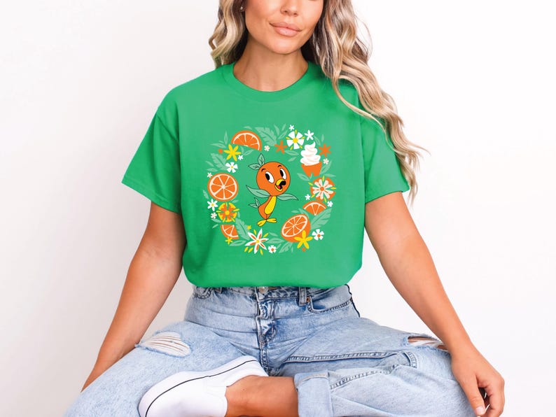 Puede incluir: Camiseta verde con un personaje de dibujos animados de naranja rodeado de rodajas de naranja, flores y postres. El dise&ntilde;o est&aacute; centrado en la camiseta. La camiseta la lleva una persona.
