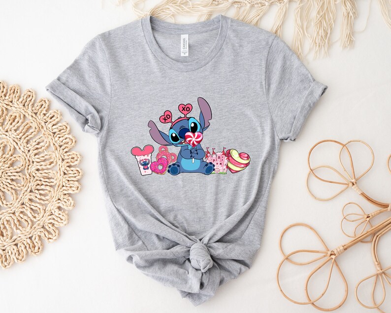 Puede incluir: Camiseta gris con un personaje azul Stitch sosteniendo una piruleta con corazones rosas y blancos. Stitch lleva una diadema con corazones rosas y las letras "XO". La camiseta tambi&eacute;n presenta un castillo rosa y blanco, una dona rosa, una cabeza de Mickey Mouse rosa y blanca y una piruleta rosa y blanca.