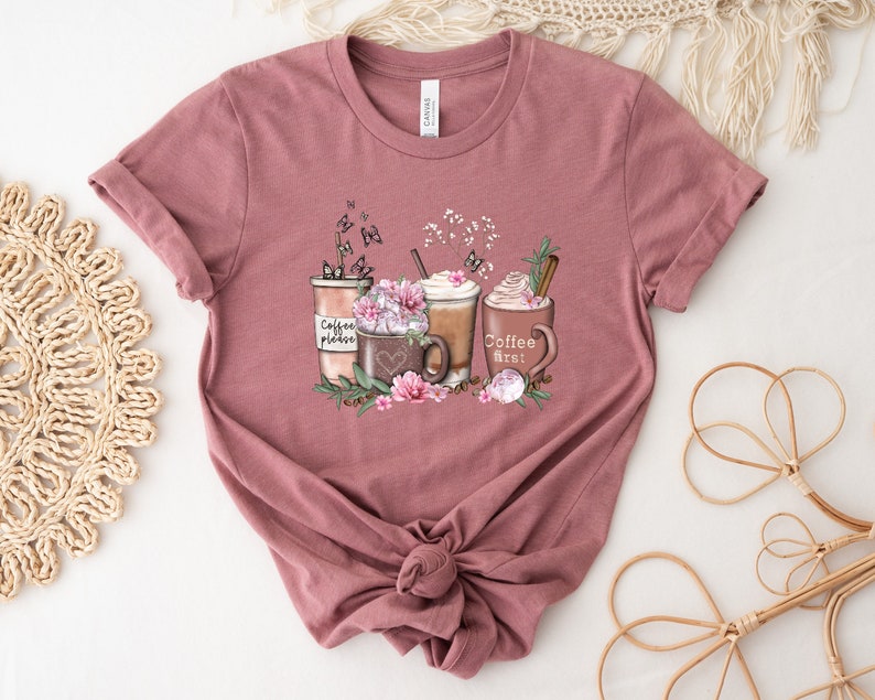 May include: A mauve t-shirt featuring a graphic of four coffee cups with whipped cream and a cinnamon stick. The cups are surrounded by flowers and butterflies. The text "Coffee First!" is on one of the cups.