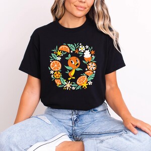 Puede incluir: Camiseta negra con un dise&ntilde;o circular que presenta un personaje de p&aacute;jaro naranja, rodajas de naranja, hojas verdes y flores blancas. El dise&ntilde;o est&aacute; centrado en la camiseta. La camiseta la lleva una persona.