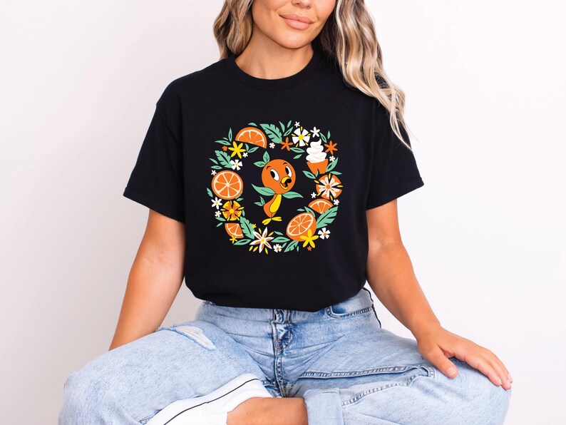Puede incluir: Camiseta negra con un dise&ntilde;o circular que presenta un personaje de p&aacute;jaro naranja, rodajas de naranja, hojas verdes y flores blancas. El dise&ntilde;o est&aacute; centrado en la camiseta. La camiseta la lleva una persona.