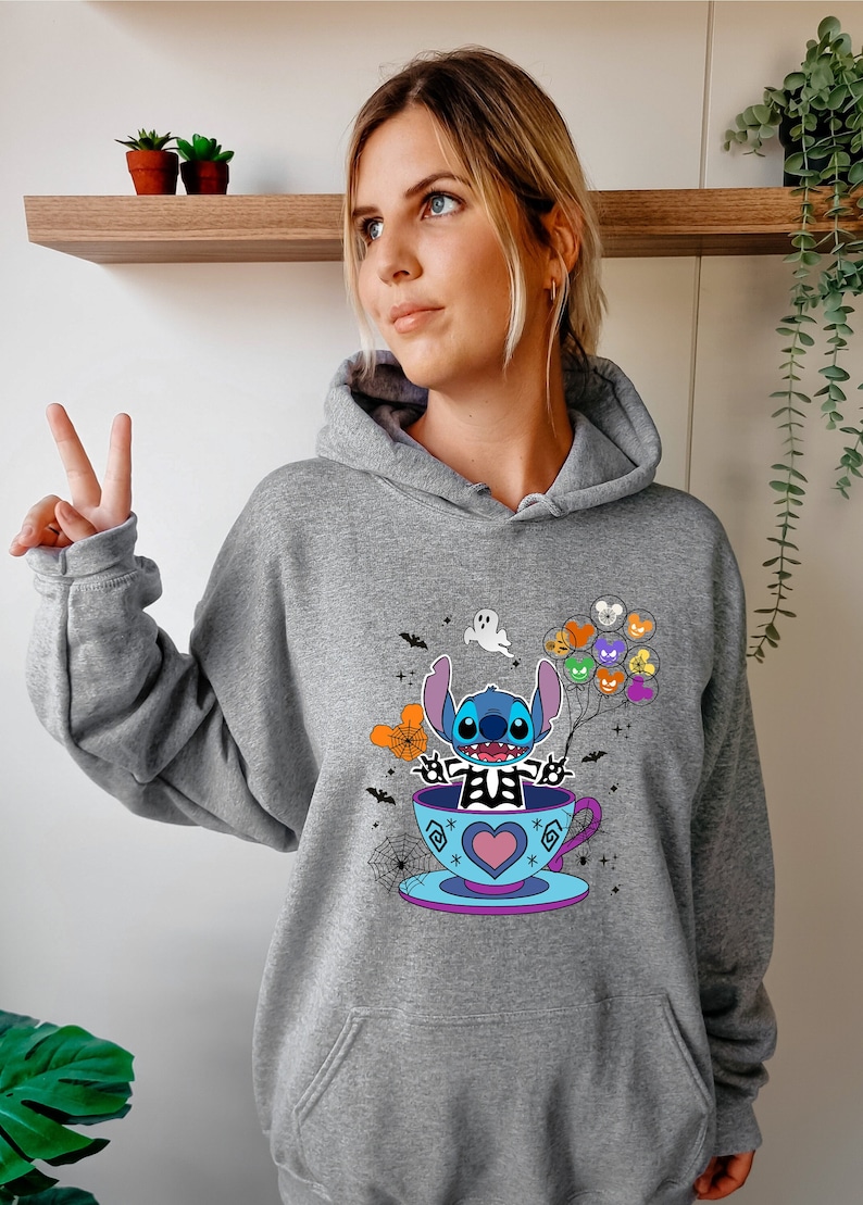 May include: Gray hooded sweatshirt with a blue Stitch character wearing a skeleton costume and holding balloons. Stitch is sitting in a teacup with a heart design. The design includes bats, spiderwebs, and a ghost.