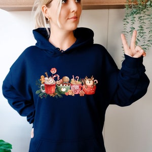 May include: A navy blue hoodie with a Christmas-themed design featuring four mugs with different holiday characters and toppings. The mugs are surrounded by festive elements like candy canes, gingerbread, and holly.
