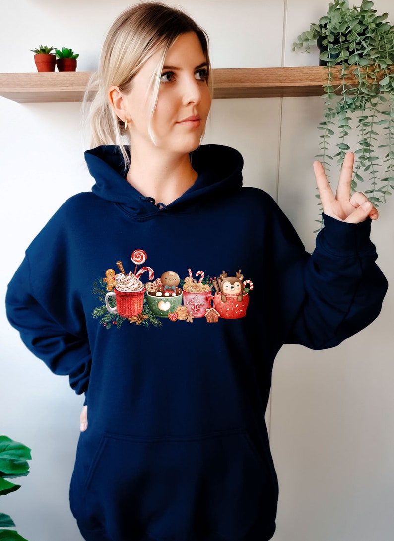 May include: A navy blue hoodie with a Christmas-themed design featuring four mugs with different holiday characters and toppings. The mugs are surrounded by festive elements like candy canes, gingerbread, and holly.