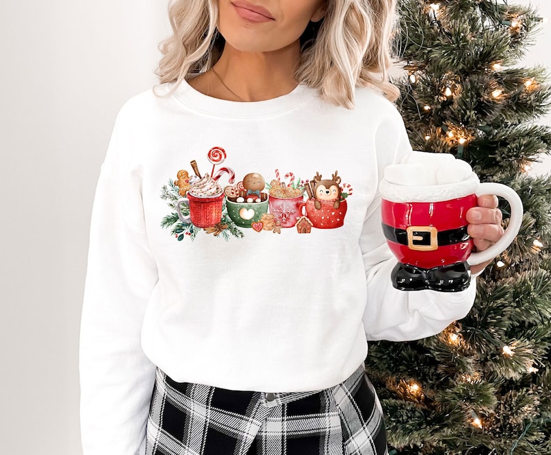 May include: White sweatshirt with a Christmas-themed design featuring four mugs of hot chocolate with whipped cream, candy canes, and gingerbread men. The mugs are decorated with red, green, and white accents.
