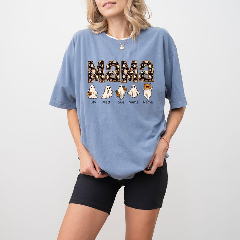 May include: A light blue t-shirt with the word "Mama" in black and white patterned letters. Five ghosts with names are lined up below the word "Mama".