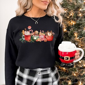 May include: Black sweatshirt with a festive design of four mugs filled with hot chocolate, marshmallows, candy canes, and gingerbread cookies. The mugs are decorated with Christmas-themed designs, including a reindeer, a snowman, and a gingerbread man.