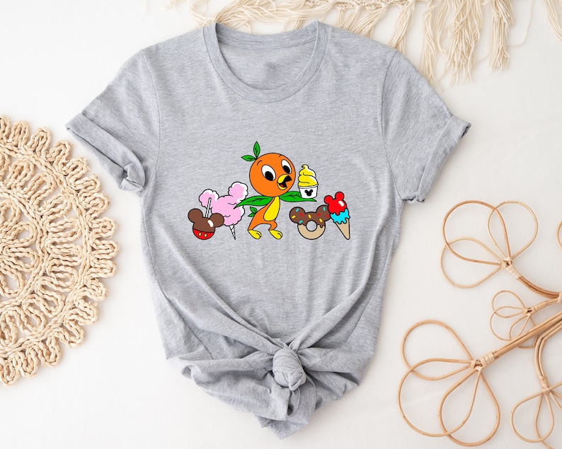 May include: A gray t-shirt with a cartoon orange bird holding a green leaf. The bird is surrounded by cartoon images of a pink cotton candy, a brown Mickey Mouse ear hat, a yellow ice cream cone, a brown donut with sprinkles, and a red and white ice cream cone.