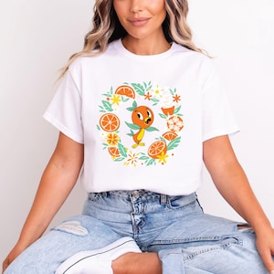 Puede incluir: Camiseta blanca con un personaje de dibujos animados de naranja rodeado de rodajas de naranja, flores y hojas. El dise&ntilde;o utiliza naranja, amarillo y verde. La camiseta tiene un cuello redondo cl&aacute;sico.