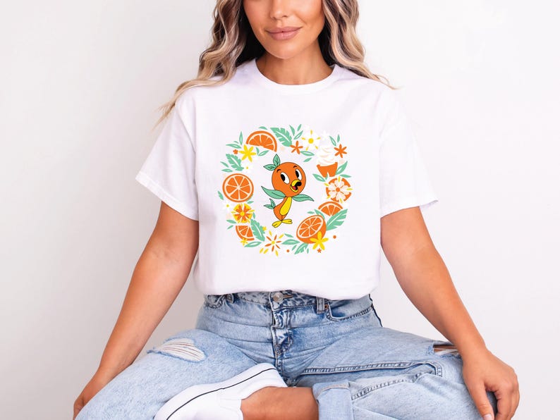 Puede incluir: Camiseta blanca con un personaje de dibujos animados de naranja rodeado de rodajas de naranja, flores y hojas. El dise&ntilde;o utiliza naranja, amarillo y verde. La camiseta tiene un cuello redondo cl&aacute;sico.