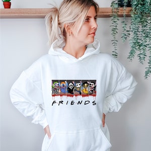 Op de afbeelding: Witte hoodie met een afbeelding van horrorfilmfiguren in een "Friends"-stijl ontwerp. De personages zijn Stitch, Ghostface, Jason Voorhees, Freddy Krueger en Michael Myers. De tekst "FRIENDS" is onder de afbeelding gedrukt.