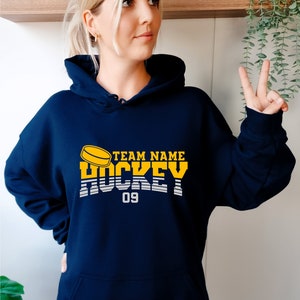 Może przedstawiać: Bordowa bluza z kapturem z ż&oacute;łto-białym nadrukiem z napisem "TEAM NAME HOCKEY 09".