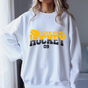 Może przedstawiać: Biała bluza z ż&oacute;łto-czarnym nadrukiem graficznym z napisem "TEAM NAME HOCKEY 09" i grafiką hokejowej krążka.