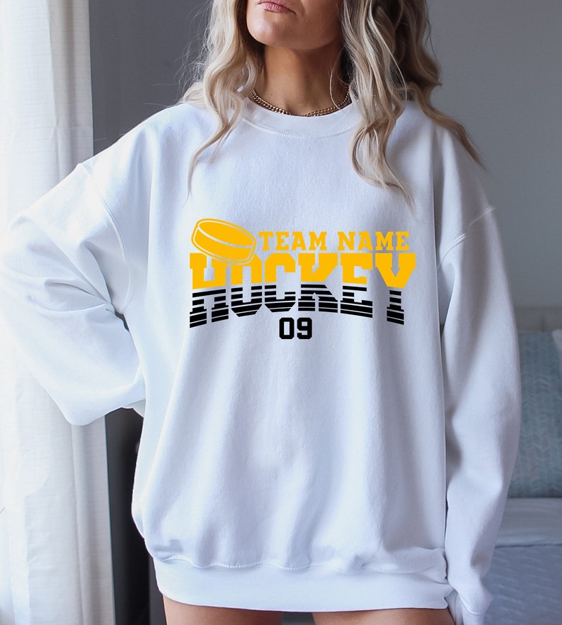 Może przedstawiać: Biała bluza z ż&oacute;łto-czarnym nadrukiem graficznym z napisem "TEAM NAME HOCKEY 09" i grafiką hokejowej krążka.