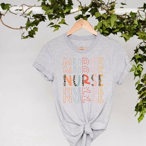 May include: A gray t-shirt with the word "NURSE" printed in colorful letters with a leopard print pattern. The letters are stacked on top of each other and are repeated multiple times.