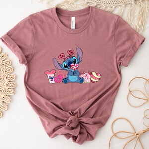 Puede incluir: Una camiseta color malva con un personaje azul Stitch que lleva una diadema rosa en forma de coraz&oacute;n y sostiene una piruleta rosa. El personaje est&aacute; rodeado de corazones rosas, una rosquilla rosa y un bast&oacute;n de caramelo rosa y blanco.