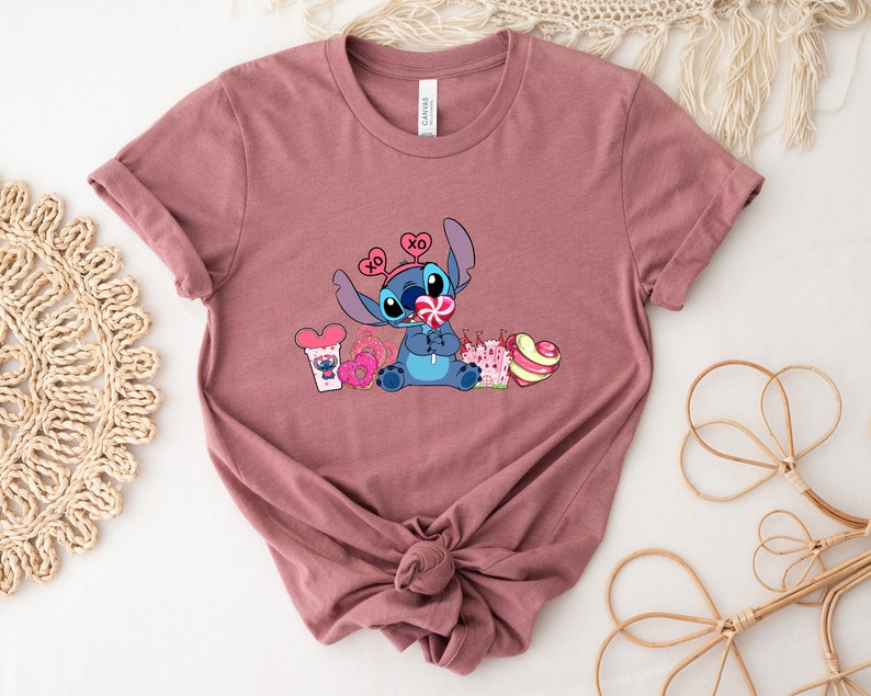 Puede incluir: Una camiseta color malva con un personaje azul Stitch que lleva una diadema rosa en forma de coraz&oacute;n y sostiene una piruleta rosa. El personaje est&aacute; rodeado de corazones rosas, una rosquilla rosa y un bast&oacute;n de caramelo rosa y blanco.