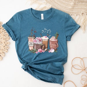 May include: Teal blue t-shirt featuring a graphic of three coffee cups with whipped cream and flowers. The text on the cups reads "Coffee please", "Coffee first", and "Coffee Street".
