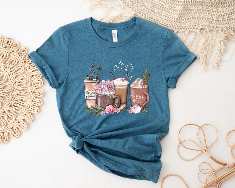 May include: Teal blue t-shirt featuring a graphic of three coffee cups with whipped cream and flowers. The text on the cups reads "Coffee please", "Coffee first", and "Coffee Street".