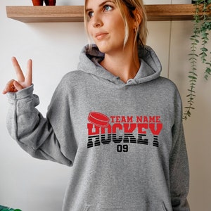 Może przedstawiać: Szara bluza z kapturem z grafiką czerwonej hokejowej krążka i tekstem "TEAM NAME HOCKEY 09".