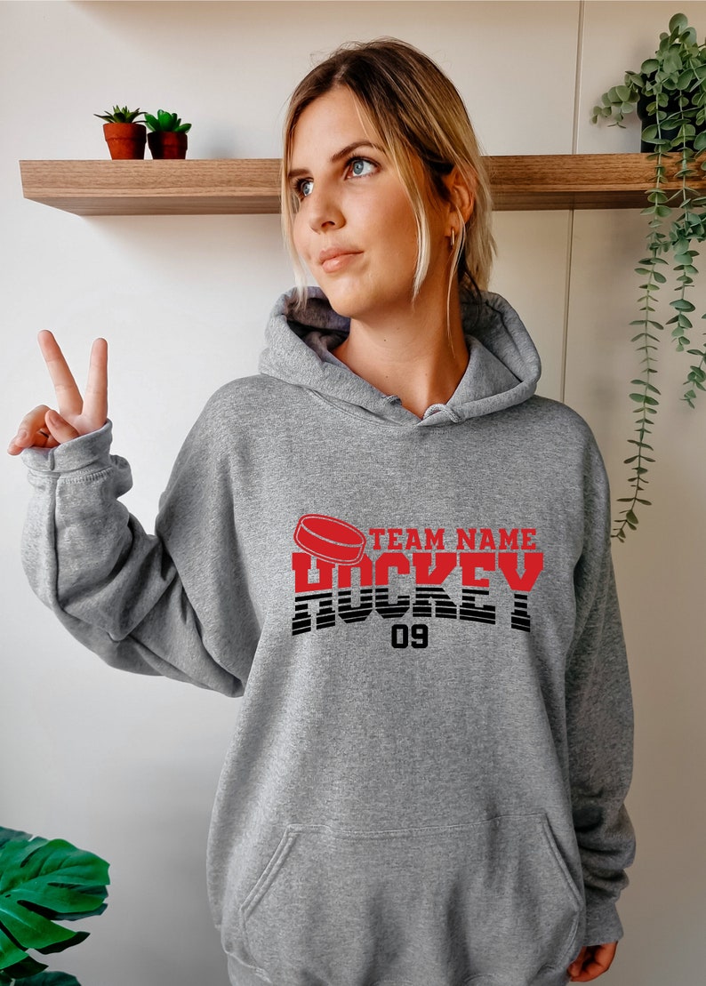 Może przedstawiać: Szara bluza z kapturem z grafiką czerwonej hokejowej krążka i tekstem "TEAM NAME HOCKEY 09".