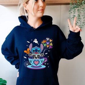 May include: A navy blue hoodie with a graphic of Stitch in a teacup holding Mickey Mouse balloons. The graphic also includes a ghost, bats, and spiderwebs.