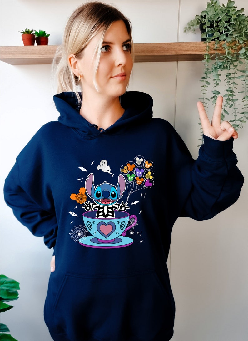 May include: A navy blue hoodie with a graphic of Stitch in a teacup holding Mickey Mouse balloons. The graphic also includes a ghost, bats, and spiderwebs.