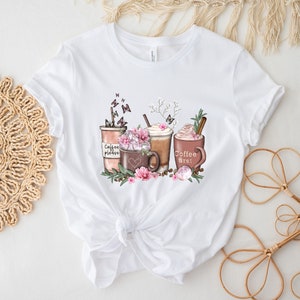 May include: White t-shirt featuring a graphic of three coffee drinks with whipped cream, flowers, and butterflies. One of the cups has the text "Coffee first!" on it.
