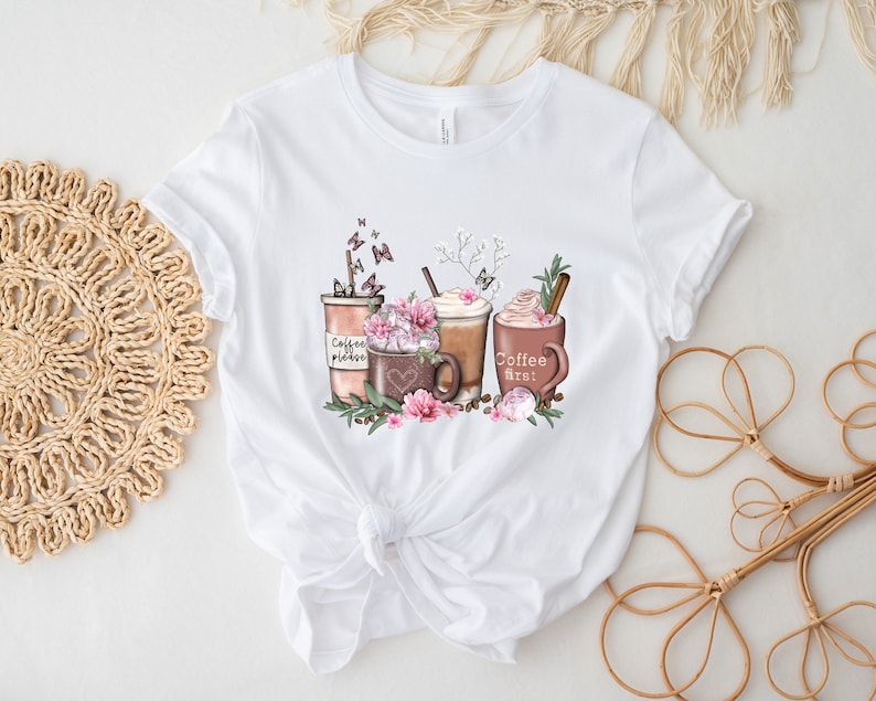 May include: White t-shirt featuring a graphic of three coffee drinks with whipped cream, flowers, and butterflies. One of the cups has the text "Coffee first!" on it.