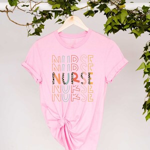 May include: Pink t-shirt with the word "NURSE" printed in colorful letters with a leopard print design. The word "NURSE" is repeated multiple times in a stacked pattern.