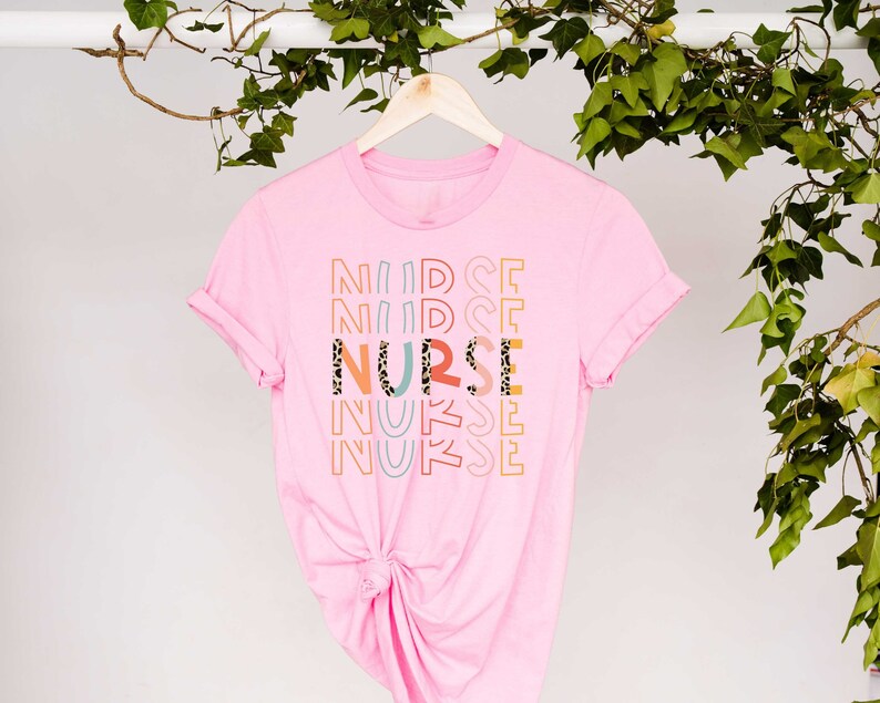 May include: Pink t-shirt with the word "NURSE" printed in colorful letters with a leopard print design. The word "NURSE" is repeated multiple times in a stacked pattern.