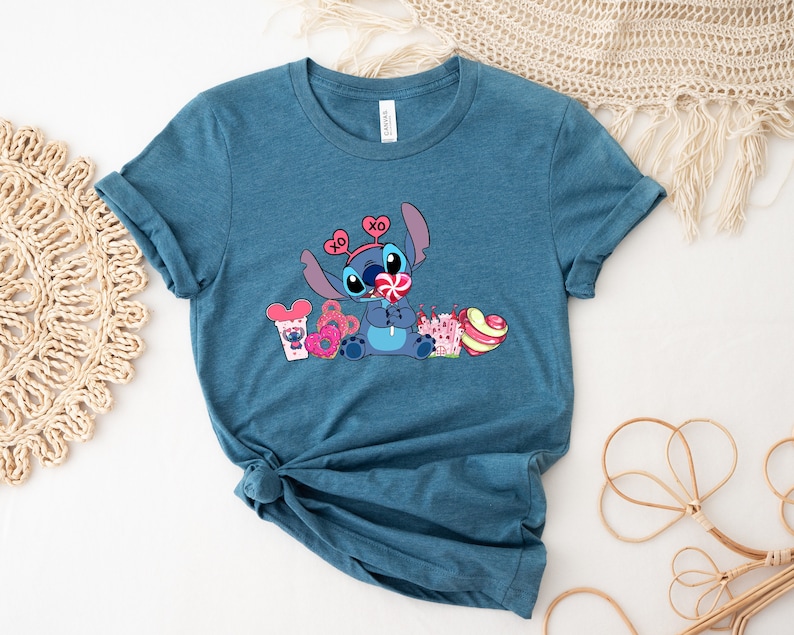 Puede incluir: Una camiseta azul turquesa con una imagen de dibujos animados de Stitch de Lilo & Stitch. Stitch lleva una diadema rosa con las letras XO y sostiene una piruleta. Est&aacute; rodeado de corazones rosas y rojos, una rosquilla rosa y una taza azul con una oreja de Mickey Mouse.