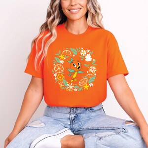 Puede incluir: Camiseta naranja brillante con un dise&ntilde;o de p&aacute;jaro naranja de dibujos animados. El p&aacute;jaro est&aacute; rodeado por una corona de flores, rodajas de naranja y hojas. El dise&ntilde;o de la camiseta es alegre y colorido.