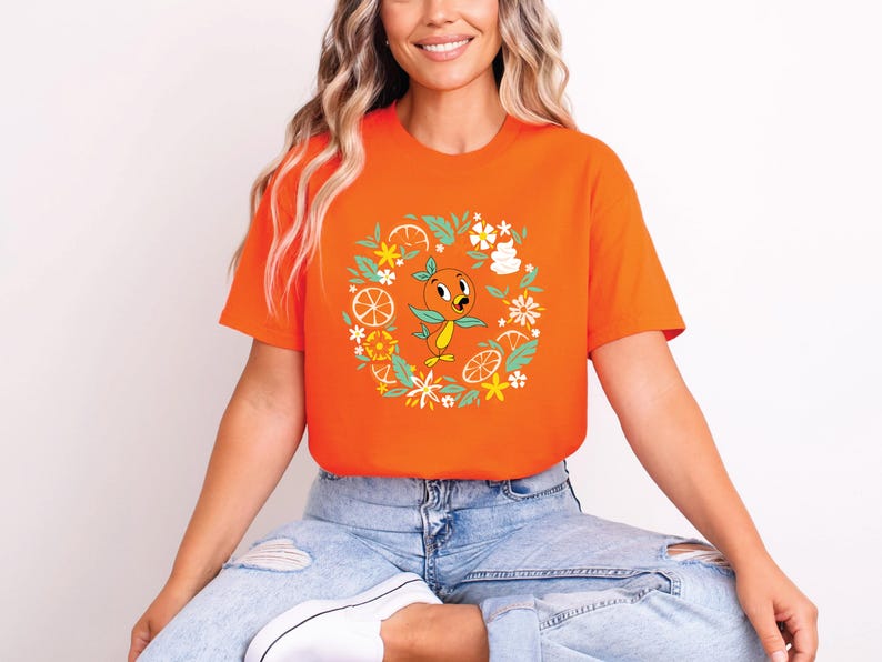 Puede incluir: Camiseta naranja brillante con un dise&ntilde;o de p&aacute;jaro naranja de dibujos animados. El p&aacute;jaro est&aacute; rodeado por una corona de flores, rodajas de naranja y hojas. El dise&ntilde;o de la camiseta es alegre y colorido.