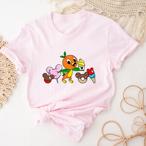 May include: Pink t-shirt with a cartoon orange bird holding a green leaf and a cup of ice cream. The bird is surrounded by other cartoon food items, including a Mickey Mouse ear ice cream cone, a donut, and cotton candy.