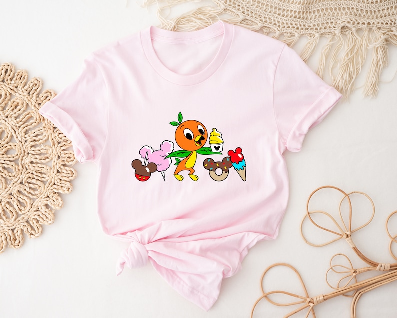 May include: Pink t-shirt with a cartoon orange bird holding a green leaf and a cup of ice cream. The bird is surrounded by other cartoon food items, including a Mickey Mouse ear ice cream cone, a donut, and cotton candy.